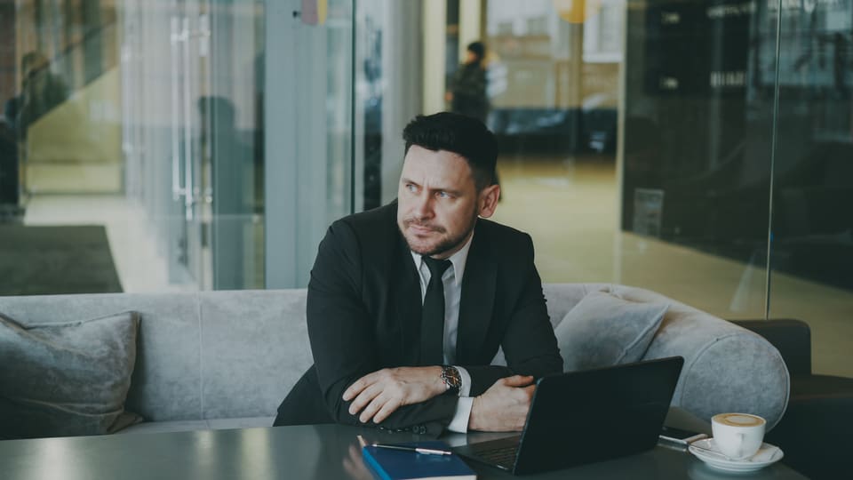 Een man in pak op een bank in een lichte lobby, met zijn laptop, die peinzend opzij kijkt