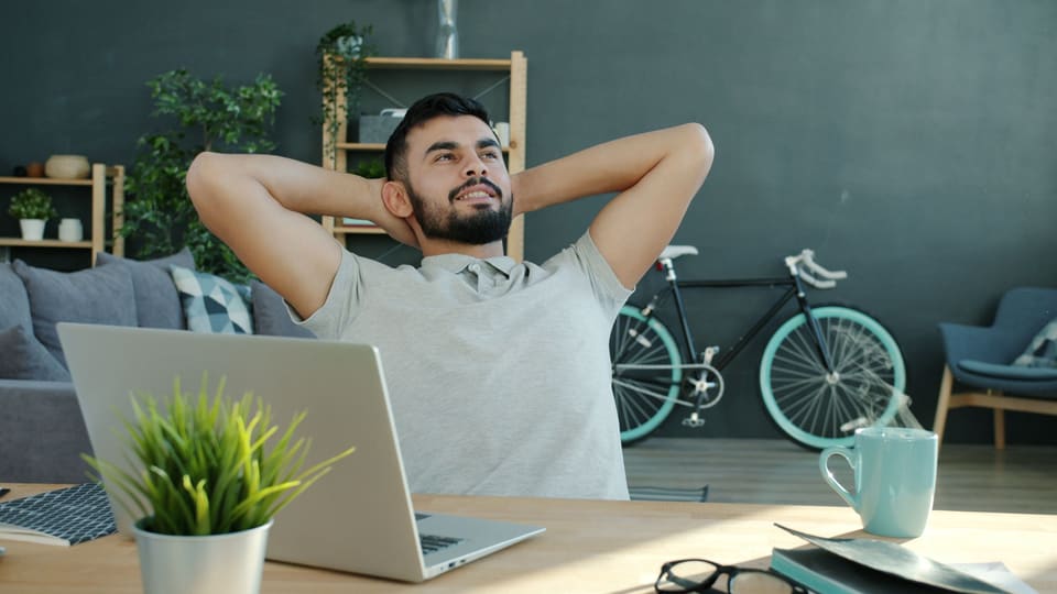 Een man die ontspannen achteroverleunt aan zijn bureau, met zijn handen achter zijn hoofd en een glimlach
